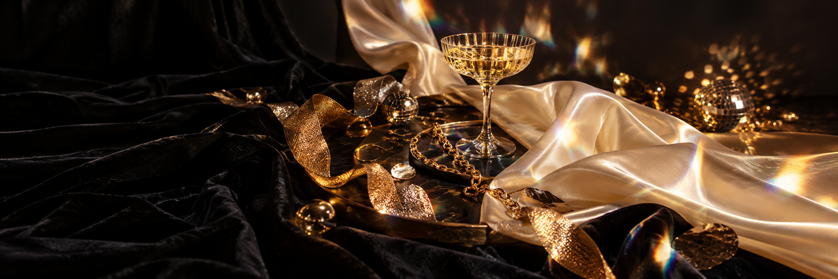 Natureza-morta editorial de festa em veludo preto e cetim marfim, com correntes douradas, taça, ornamentos espelhados e reflexos prismáticos